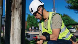 A field worker wearing safety gear inspects a utility pole, using a handheld device in a sunny outdoor setting. - Agentic AI for field operations